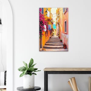 May include: A vertical wall art print depicting a sunlit Mediterranean street scene with stone stairs leading toward the sea. Vibrant pink bougainvillea flowers drape over yellow walls, while laundry hangs on lines and potted plants line the steps.