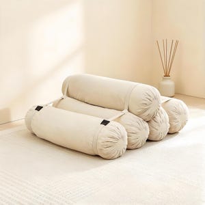 May include: A set of beige cylindrical pillows arranged to resemble a chair. The pillows are made of a soft fabric and are connected with straps. The scene is set on a cream-colored rug, with a vase and reeds in the background.