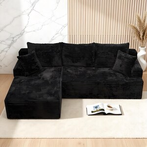 Op de afbeelding: Een zwarte hoekbank van corduroy met twee bijpassende kussens. De bank heeft een chaise longue en staat op een wit tapijt. Een boek en een kopje koffie liggen op het tapijt voor de bank. De achtergrond heeft een marmeren muur en een houten paneel.