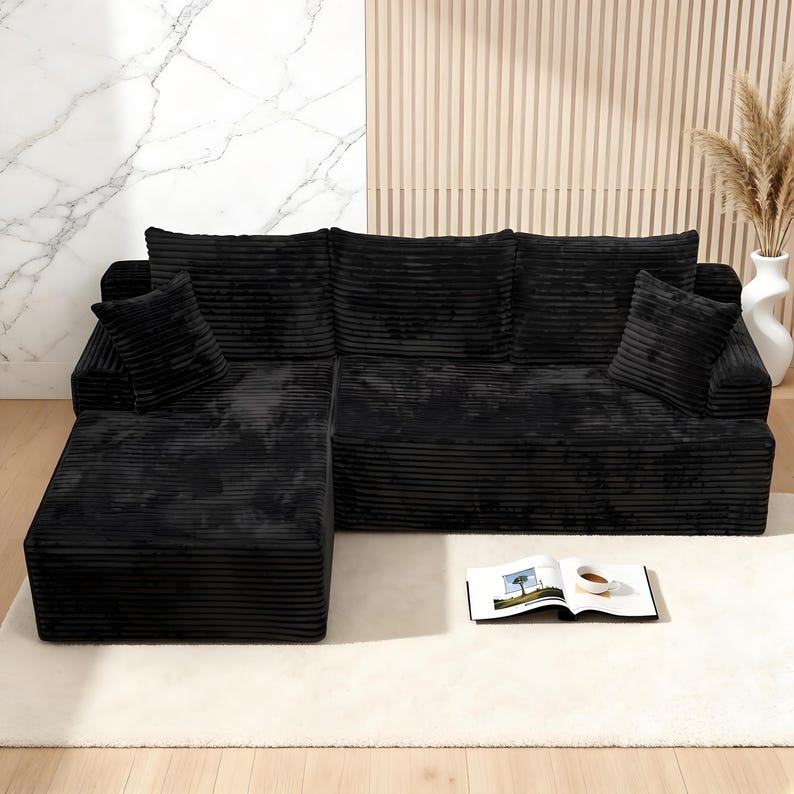 Op de afbeelding: Een zwarte hoekbank van corduroy met twee bijpassende kussens. De bank heeft een chaise longue en staat op een wit tapijt. Een boek en een kopje koffie liggen op het tapijt voor de bank. De achtergrond heeft een marmeren muur en een houten paneel.