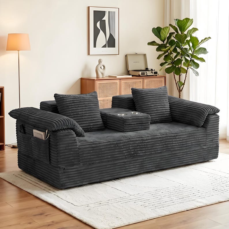 Może przedstawiać: Ciemnoszara sofa sztruksowa o teksturowanej, prążkowanej tkaninie, z dwiema pasującymi kwadratowymi poduszkami i składaną konsolą środkową z dwoma wbudowanymi uchwytami na kubki. Boczny podłokietnik posiada wbudowaną kieszeń do przechowywania.