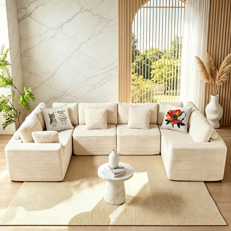 May include: Cream-colored modular sectional sofa with corduroy texture. Includes multiple square and rectangular throw pillows. A round marble side table and a beige rug complete the living room setting. Decorative pillows feature a cat and a floral design.