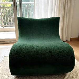 May include: A dark green, modern armchair with a curved, organic shape. The chair is upholstered in a textured fabric, and it sits on a neutral-colored rug. The chair's design is minimalist and contemporary, suitable for a living room or lounge area.