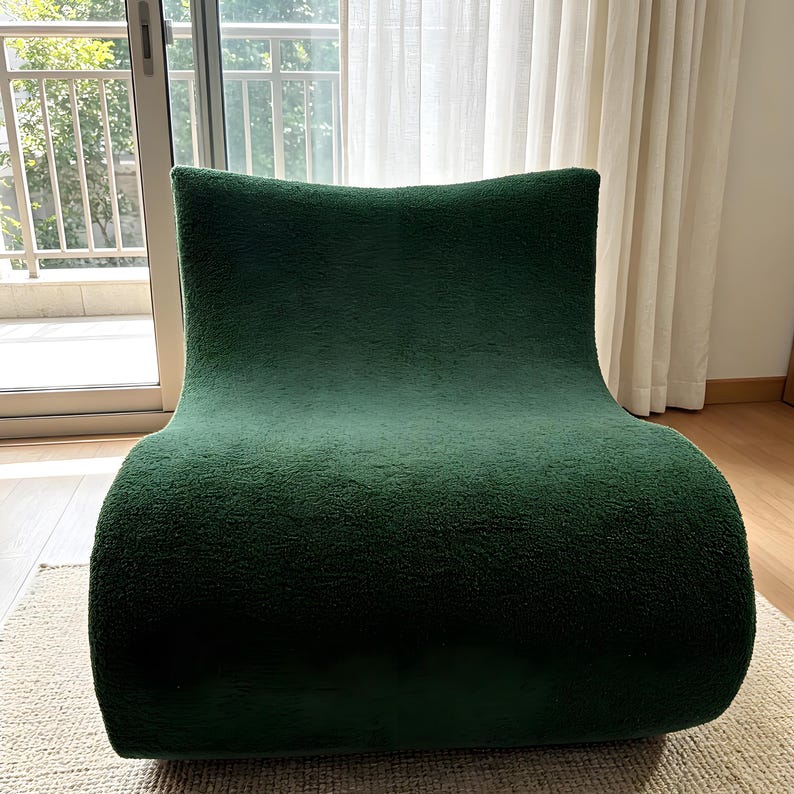 May include: A dark green, modern armchair with a curved, organic shape. The chair is upholstered in a textured fabric, and it sits on a neutral-colored rug. The chair's design is minimalist and contemporary, suitable for a living room or lounge area.