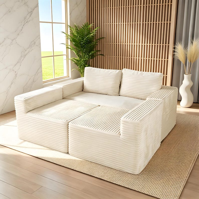 May include: A modular, cream-colored sectional sofa with a corduroy texture. The sofa is composed of several square and rectangular pieces, including armrests and cushions. The sofa sits on a beige rug in a well-lit room with a window and decorative plants.