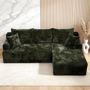 Op de afbeelding: Een donkergroene corduroy hoekbank met bijpassende kussens. De bank heeft een modern design en staat op een wit tapijt. De achtergrond heeft een marmeren muur en een houten paneelmuur.