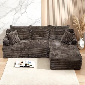 Op de afbeelding: Een hoekbank van donkergrijze corduroy stof. De bank heeft een chaise longue en twee bijpassende sierkussens. Voor de bank ligt een wit hoogpolig tapijt, met een boek en een kopje koffie.