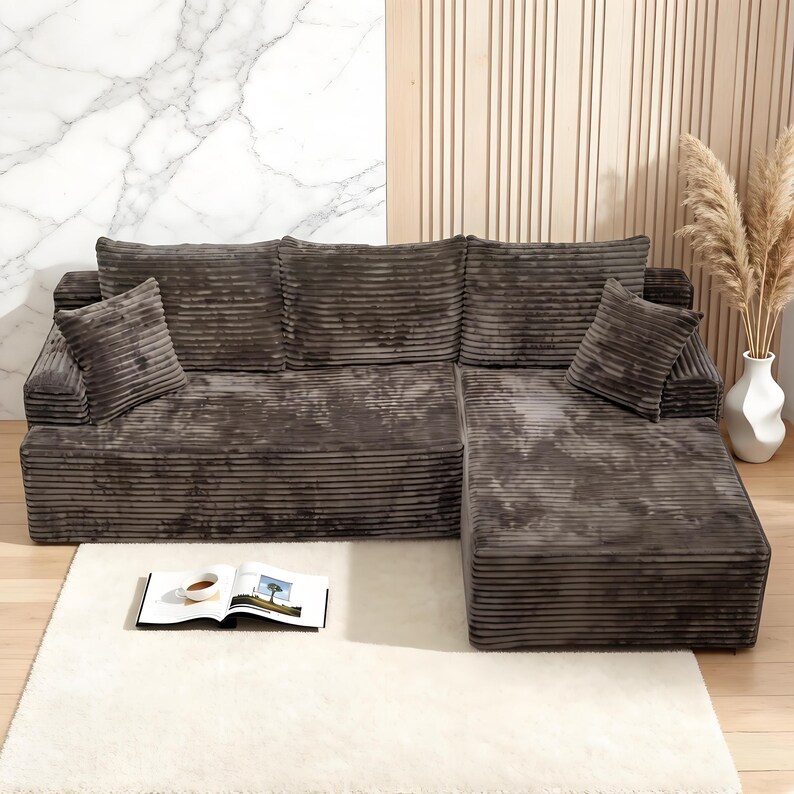 Op de afbeelding: Een hoekbank van donkergrijze corduroy stof. De bank heeft een chaise longue en twee bijpassende sierkussens. Voor de bank ligt een wit hoogpolig tapijt, met een boek en een kopje koffie.