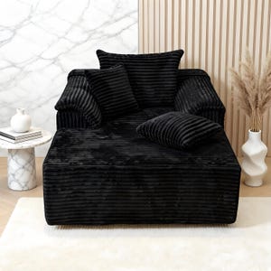 May include: A black corduroy-style lounge chair with matching cushions. The chair has a low profile and a plush appearance. The cushions are arranged on the seat and back, creating a comfortable seating area. The chair sits on a white rug.