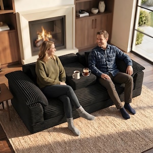 Może przedstawiać: Czarna sofa sztruksowa z konsolą środkową wyposażoną w dwa uchwyty na kubki. Sofa ma teksturowane, prążkowane wykończenie tkaniny, szerokie podłokietniki i głębokie siedzisko. Mebel stoi na jasnym, plecionym dywanie w salonie.