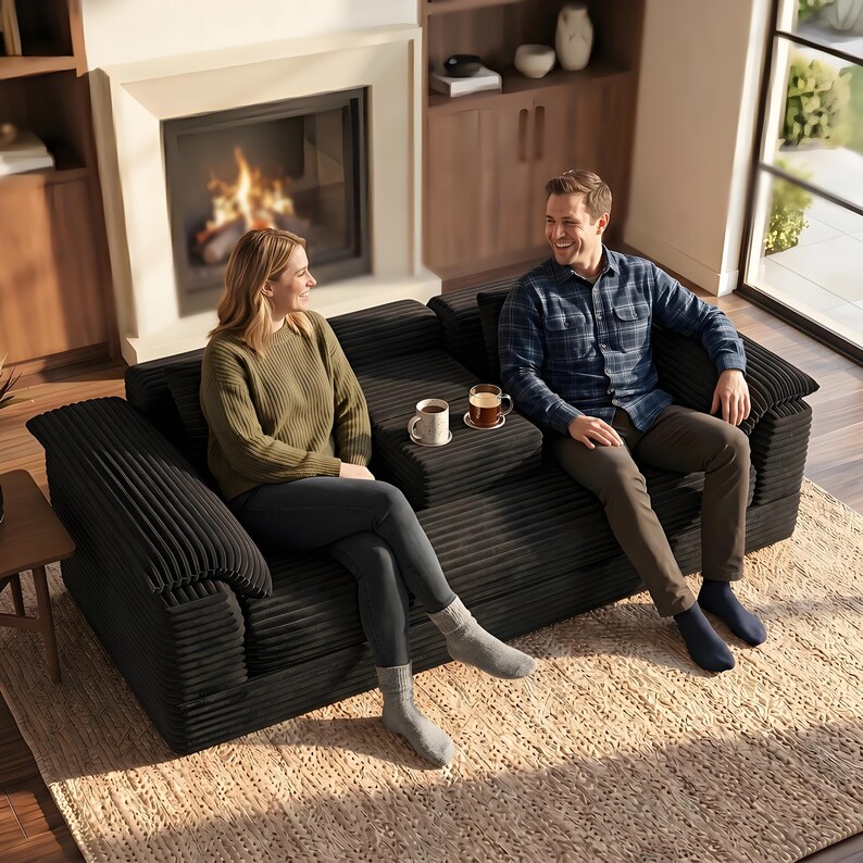 Może przedstawiać: Czarna sofa sztruksowa z konsolą środkową wyposażoną w dwa uchwyty na kubki. Sofa ma teksturowane, prążkowane wykończenie tkaniny, szerokie podłokietniki i głębokie siedzisko. Mebel stoi na jasnym, plecionym dywanie w salonie.