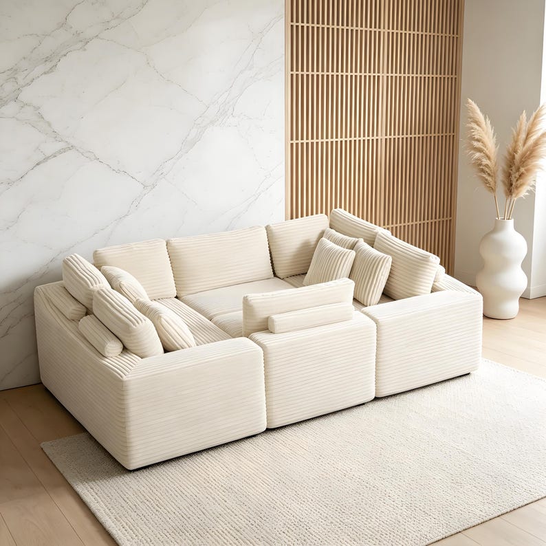 May include: A modular, cream-colored corduroy sectional sofa with multiple cushions. The sofa is arranged in an L-shape and sits on a light-colored rug. The background includes a marble wall, a wooden slat screen, and a white vase with decorative dried plants.