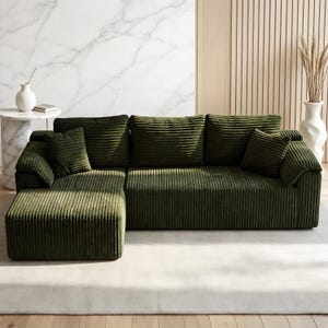 May include: A large, olive green corduroy sectional sofa with multiple cushions. The sofa has a chaise lounge on the left side and is placed on a light-colored rug. A white vase with dried plants is visible in the background.