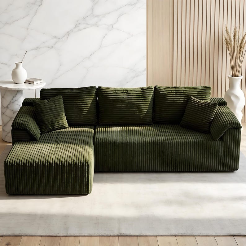 May include: A large, olive green corduroy sectional sofa with multiple cushions. The sofa has a chaise lounge on the left side and is placed on a light-colored rug. A white vase with dried plants is visible in the background.