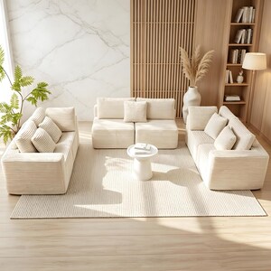 May include: A modern living room set featuring a cream-colored corduroy sofa, loveseat, and armchair with matching throw pillows. A small, round white side table sits on a cream-colored rug. The room has a neutral color palette with wooden accents.