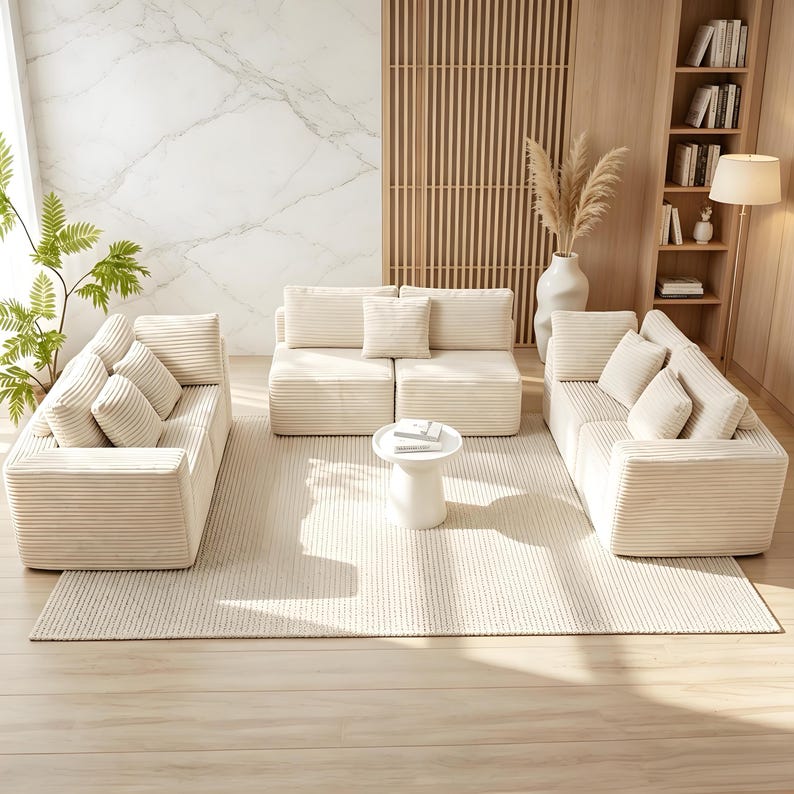 May include: A modern living room set featuring a cream-colored corduroy sofa, loveseat, and armchair with matching throw pillows. A small, round white side table sits on a cream-colored rug. The room has a neutral color palette with wooden accents.
