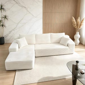 May include: A large, off-white sectional sofa with a corduroy texture. The sofa has a chaise lounge on the left side and several matching throw pillows. The room features a marble accent wall, wooden paneling, and a neutral-colored rug.