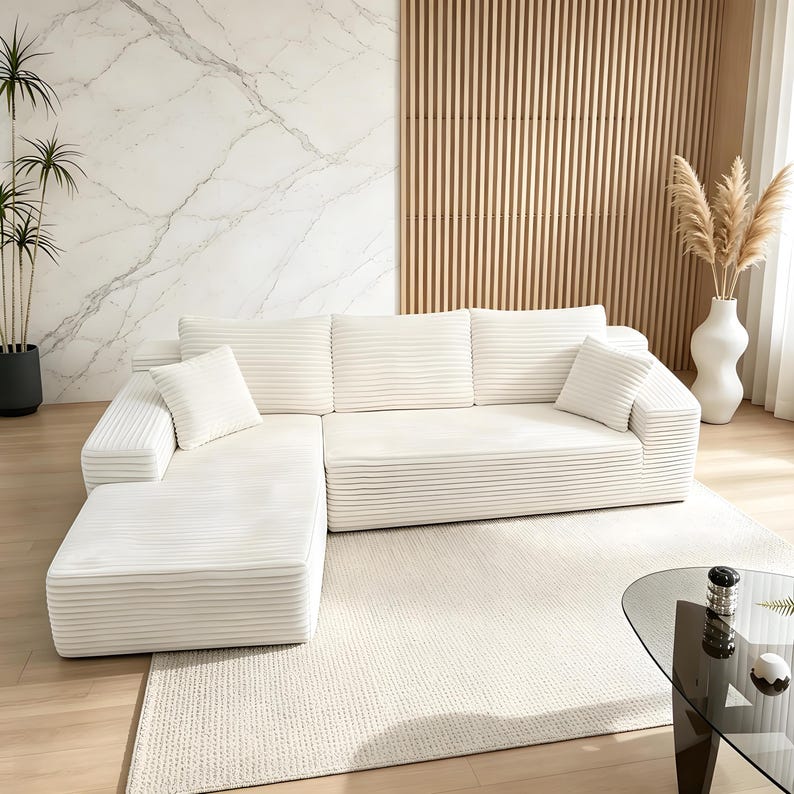 May include: A large, off-white sectional sofa with a corduroy texture. The sofa has a chaise lounge on the left side and several matching throw pillows. The room features a marble accent wall, wooden paneling, and a neutral-colored rug.