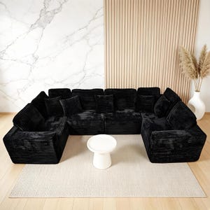 May include: A large, U-shaped black corduroy sectional sofa with multiple cushions. A small, white, round side table sits in front of the sofa on a beige rug. The background features a marble wall and a wooden slatted wall.