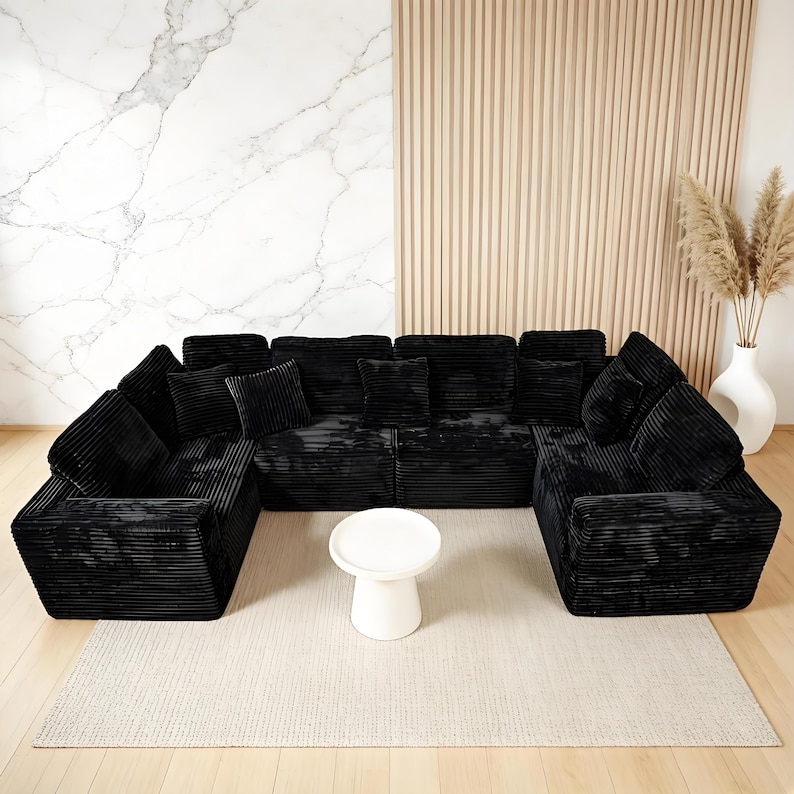 May include: A large, U-shaped black corduroy sectional sofa with multiple cushions. A small, white, round side table sits in front of the sofa on a beige rug. The background features a marble wall and a wooden slatted wall.