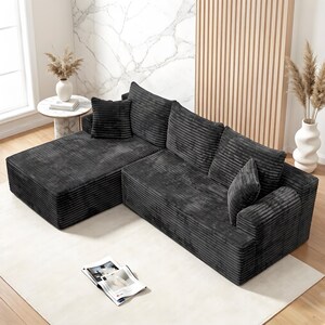 Op de afbeelding: Een donkergrijze hoekbank met een ribfluwelen textuur en een chaise longue. De bank heeft meerdere kussens en staat op een lichtgekleurd tapijt. Een tijdschrift ligt open op de vloer. Decoratieve vazen en een kleine bijzettafel zijn ook zichtbaar.