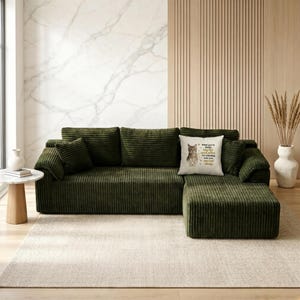 May include: A large, olive green corduroy sectional sofa with matching throw pillows. A decorative pillow with a cat illustration and text sits on the sofa. The sofa is on a neutral rug.