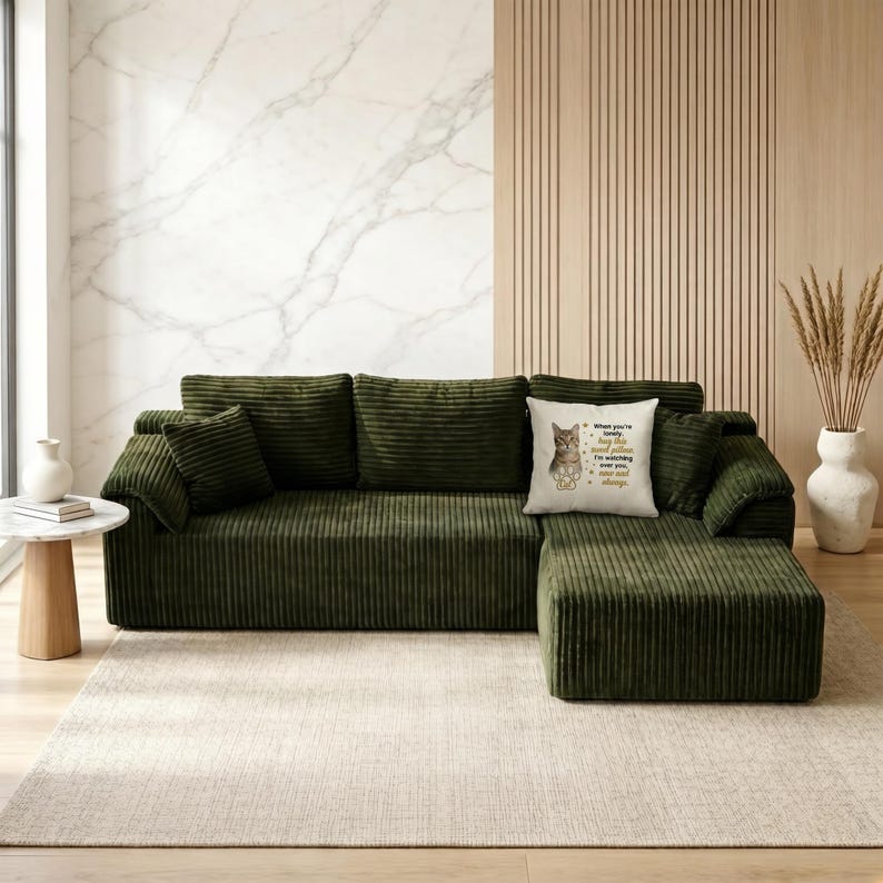 May include: A large, olive green corduroy sectional sofa with matching throw pillows. A decorative pillow with a cat illustration and text sits on the sofa. The sofa is on a neutral rug.