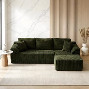 May include: An olive green corduroy sectional sofa with multiple cushions. The sofa has an L-shape design and sits on a light-colored rug. A small side table with a white marble top and a vase with dried plants are visible.