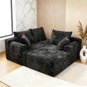 Op de afbeelding: Een modulaire, donkergrijze corduroy hoekbank met meerdere kussens en een chaise longue. De bank staat op een wit tapijt, met een vaas met gedroogde planten in de buurt. De kamer heeft een modern design met houten panelen.