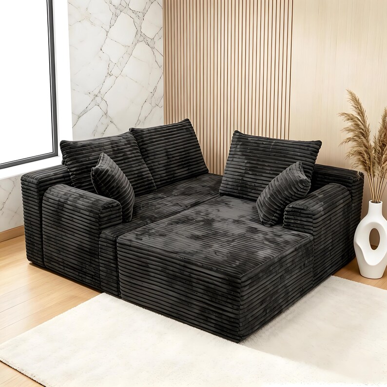 Op de afbeelding: Een modulaire, donkergrijze corduroy hoekbank met meerdere kussens en een chaise longue. De bank staat op een wit tapijt, met een vaas met gedroogde planten in de buurt. De kamer heeft een modern design met houten panelen.