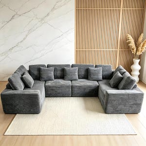 May include: A large, modular gray corduroy sofa with multiple cushions arranged on a cream-colored rug. The sofa is U-shaped and has a modern design. The background features a marble wall, wooden paneling, and a white vase with dried plants.