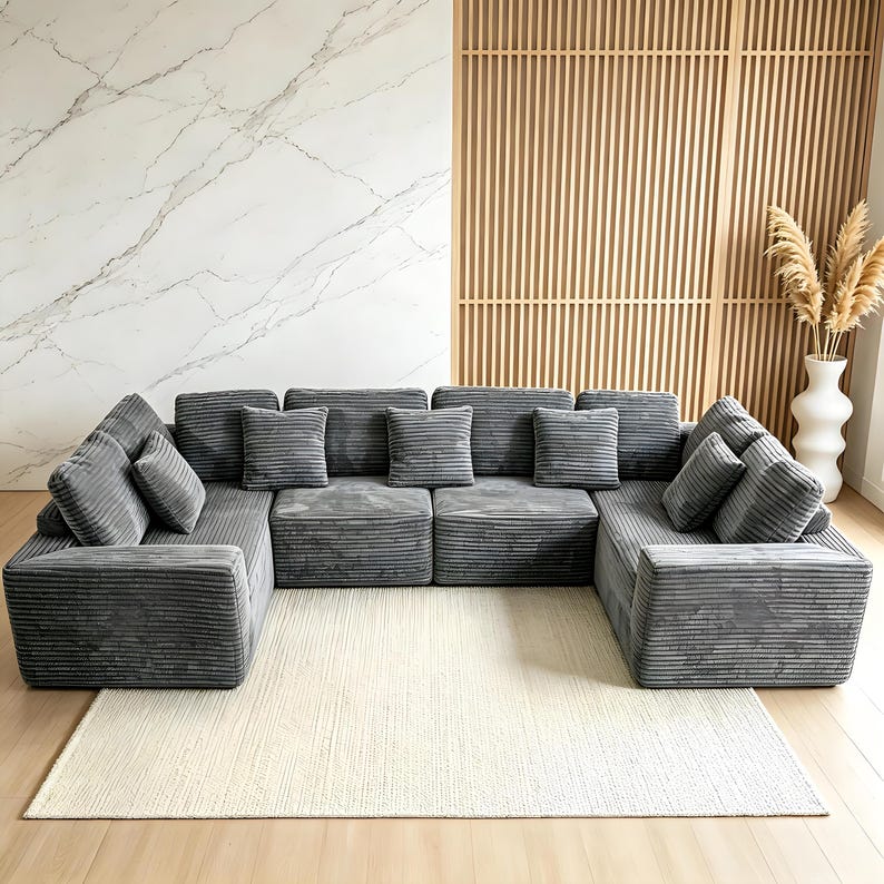May include: A large, modular gray corduroy sofa with multiple cushions arranged on a cream-colored rug. The sofa is U-shaped and has a modern design. The background features a marble wall, wooden paneling, and a white vase with dried plants.