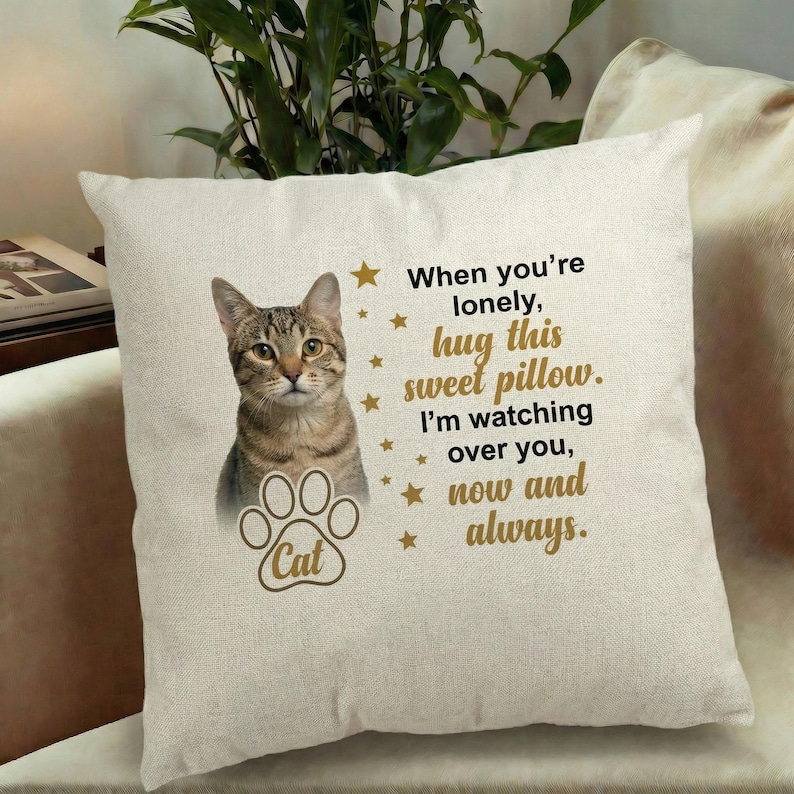 Pode incluir: Uma almofada quadrada em tom off-white com a ilustra&ccedil;&atilde;o de um gato malhado castanho, um &iacute;cone de pata com a palavra Cat e texto dourado: When you're lonely, hug this sweet pillow. I'm watching over you, now and always.