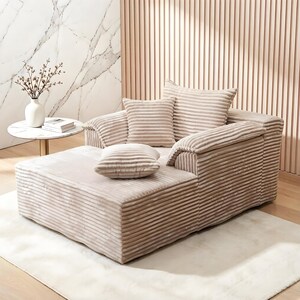 May include: A beige corduroy sectional sofa with matching ottoman and cushions. The sofa has a ribbed texture and sits on a white rug. A small side table with a vase and book is in the background.