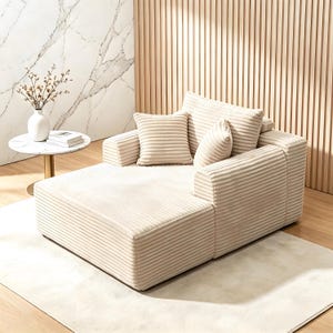 May include: Beige corduroy sectional sofa with multiple cushions. The sofa has a chaise lounge and is placed on a light-colored rug. A small round table with a white top and a vase of flowers is to the left.