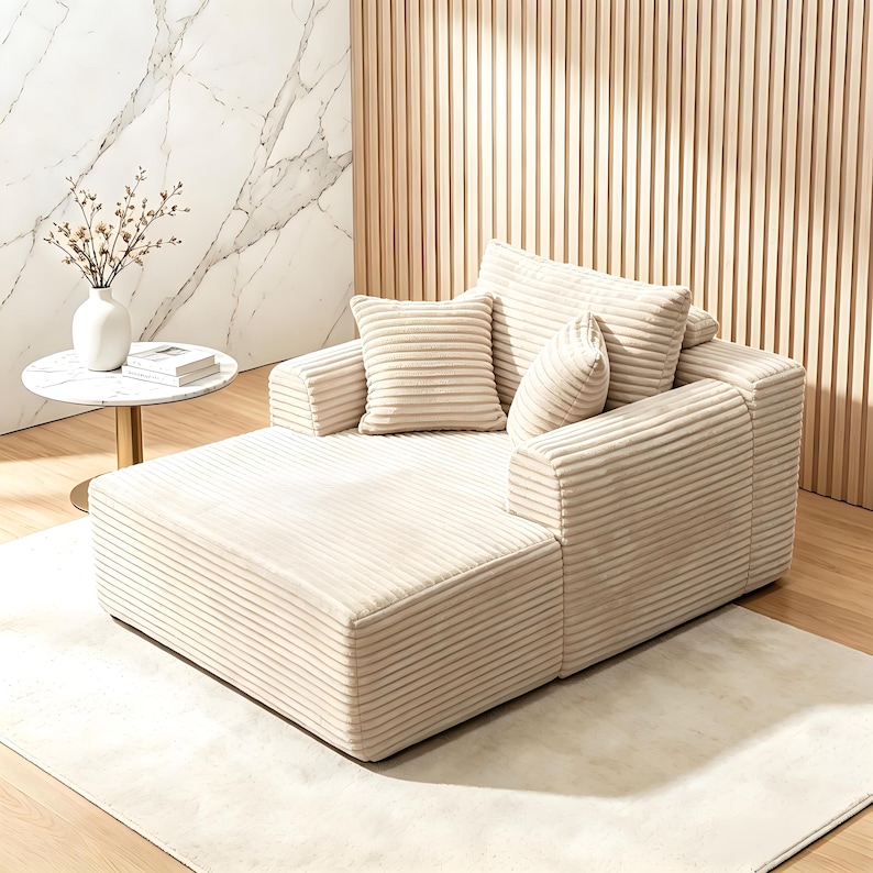 May include: Beige corduroy sectional sofa with multiple cushions. The sofa has a chaise lounge and is placed on a light-colored rug. A small round table with a white top and a vase of flowers is to the left.