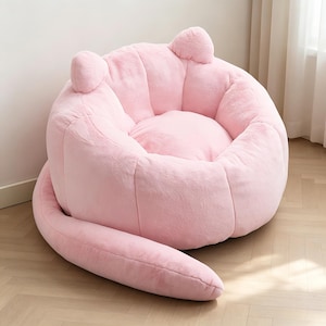 May include: A plush, pink beanbag chair with a cat-like design. The chair features two ear-shaped protrusions at the top and a long, tail-like extension. The soft, round seat is designed for comfort and relaxation.