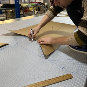 May include: A person is tracing a pattern onto light grey fabric with a pen. The fabric has a subtle texture. A brown paper pattern is placed on top of the fabric. A wooden ruler is visible in the foreground.