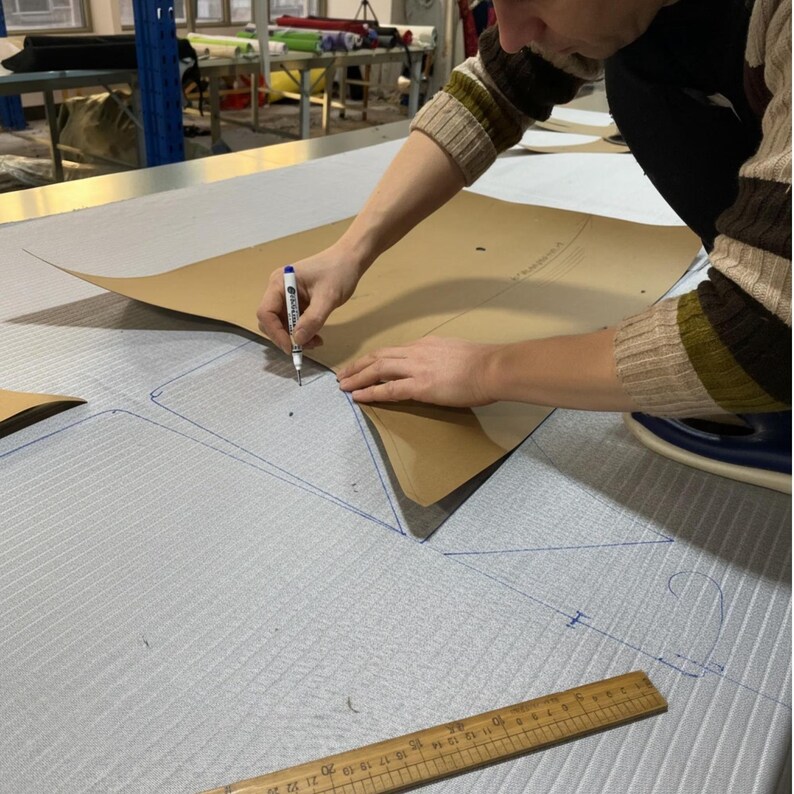 May include: A person is tracing a pattern onto light grey fabric with a pen. The fabric has a subtle texture. A brown paper pattern is placed on top of the fabric. A wooden ruler is visible in the foreground.