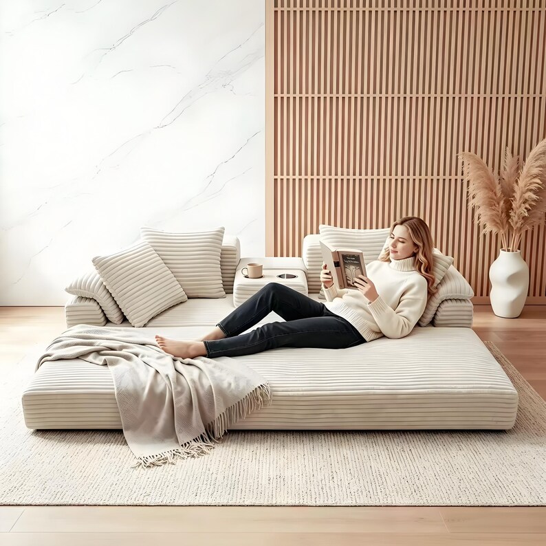 Może przedstawiać: Niska sofa podłogowa z kremowego sztruksu z wbudowaną konsolą środkową z uchwytem na kubek, kilkoma pasującymi prążkowanymi poduszkami dekoracyjnymi oraz narzuconym beżowym kocem, zapewniająca szeroką i płaską powierzchnię do siedzenia.