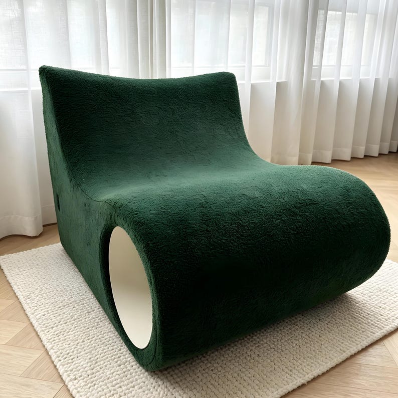 May include: A modern, emerald green lounge chair with a unique, curved design. The chair features a soft, textured fabric and a circular opening on the side. It sits on a cream-colored rug, with a white curtain in the background.