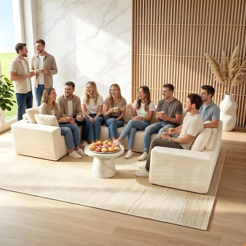 May include: A group of people are gathered in a well-lit living room, some seated on cream-colored corduroy sofas and others standing. A small marble table holds a plate of colorful pastries. The room features a large window and a wooden slatted wall.
