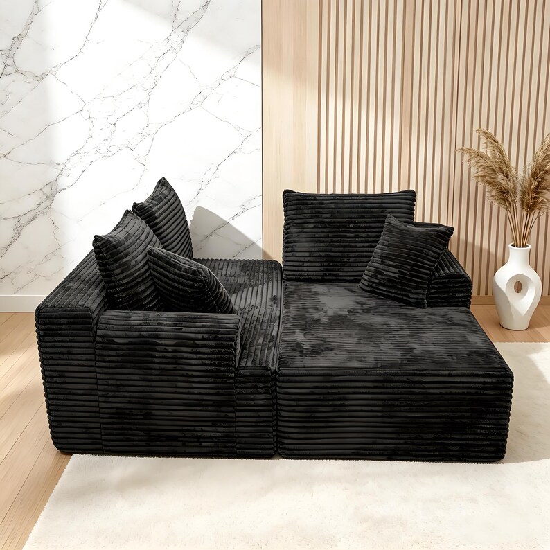 Op de afbeelding: Zwarte modulaire hoekbank met corduroy textuur en meerdere kussens. De bank is in een L-vorm gerangschikt en staat op een wit tapijt. De achtergrond heeft een marmeren muur en een houten paneelmuur.