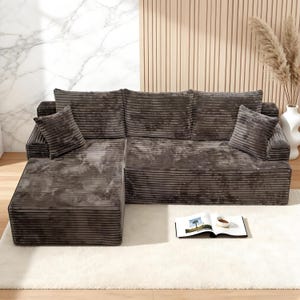 Op de afbeelding: Een hoekbank met chaise longue, bekleed met donkergrijze corduroy stof. De bank heeft twee vierkante kussens en staat op een witte shaggy vloerkleed. Een boek en een kopje koffie liggen op het vloerkleed.