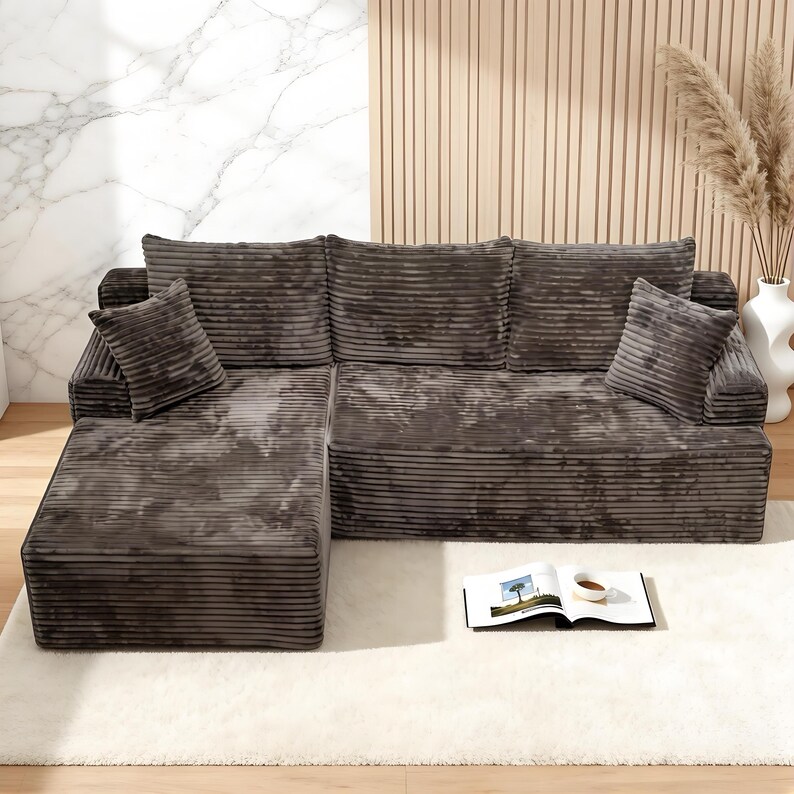 Op de afbeelding: Een hoekbank met chaise longue, bekleed met donkergrijze corduroy stof. De bank heeft twee vierkante kussens en staat op een witte shaggy vloerkleed. Een boek en een kopje koffie liggen op het vloerkleed.