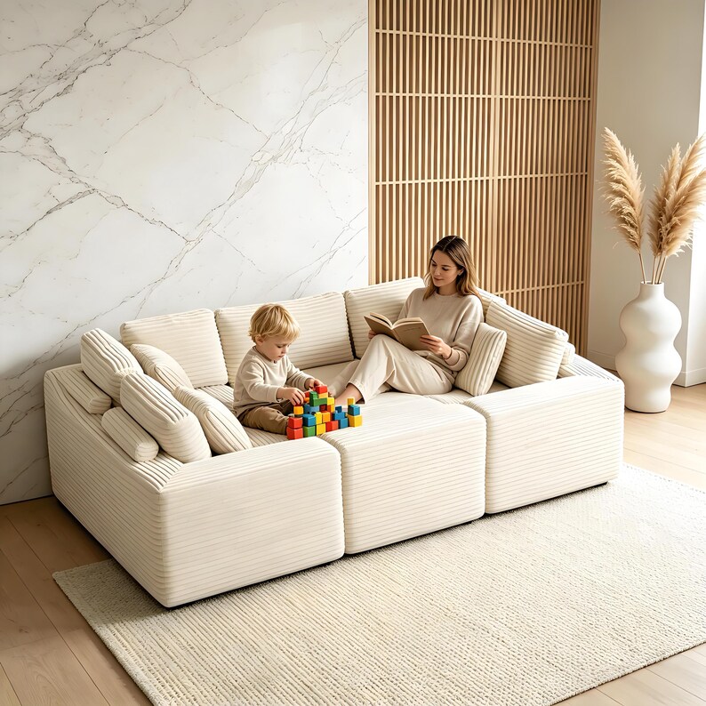 May include: A modular cream-colored corduroy sofa with multiple cushions. A woman reads a book while a child plays with colorful blocks. The sofa sits on a cream-colored rug, with a marble wall and wooden slat screen in the background.