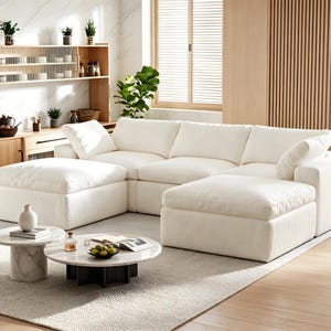 Pode incluir: Um sof&aacute; modular grande de cor branca com almofadas profundas e dois pufes a condizer. &Agrave; frente do sof&aacute; encontram-se duas mesas de centro redondas com tampos de m&aacute;rmore branco e bases cil&iacute;ndricas, uma com um vaso branco e outra com uma ta&ccedil;a de fruta verde.