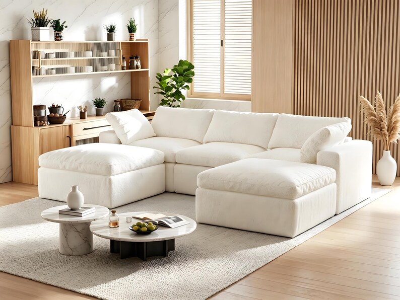Pode incluir: Um sof&aacute; modular grande de cor branca com almofadas profundas e dois pufes a condizer. &Agrave; frente do sof&aacute; encontram-se duas mesas de centro redondas com tampos de m&aacute;rmore branco e bases cil&iacute;ndricas, uma com um vaso branco e outra com uma ta&ccedil;a de fruta verde.