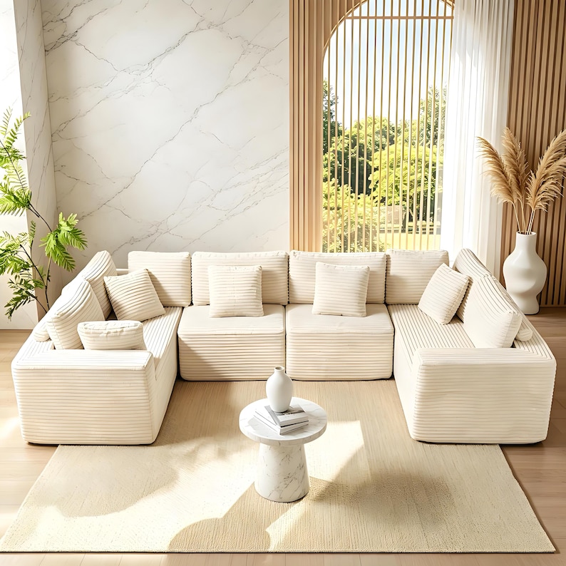 May include: A modular, cream-colored corduroy sectional sofa with multiple square cushions and throw pillows. The sofa is arranged in a U-shape on a cream rug. A small marble table and vase are in front of the sofa.
