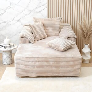 May include: A beige, corduroy-textured chaise longue with matching cushions. The longue has a low profile and is accompanied by a small side table and a decorative vase with dried plants. The background features a marble wall and a wooden panel.